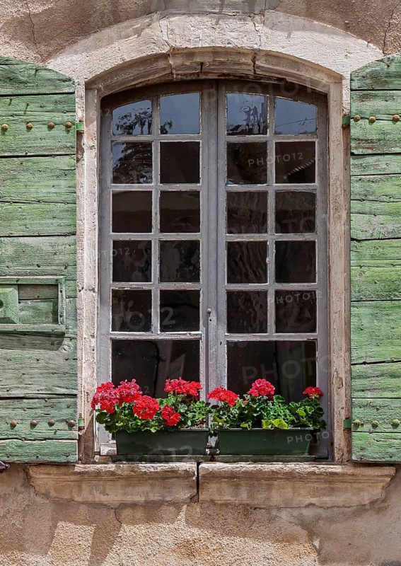 Old window with weathered green wooden shutters and red geraniums on a rustic wall, Provence, France