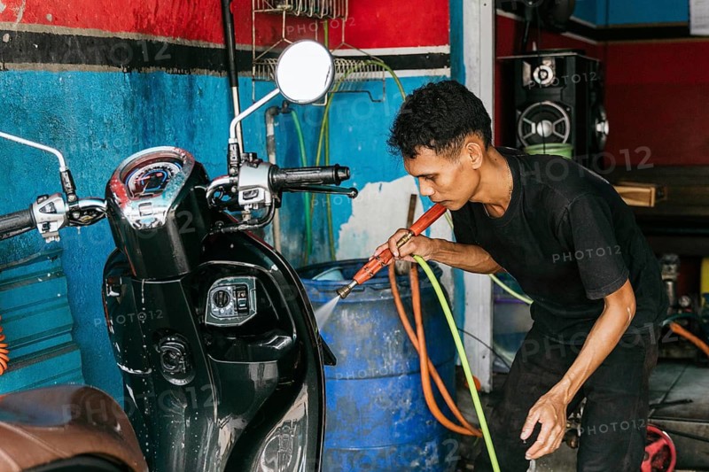 A man diligently cleans a motorcycle using a hose in a garage setting. The background features colorful walls and cleaning equipment