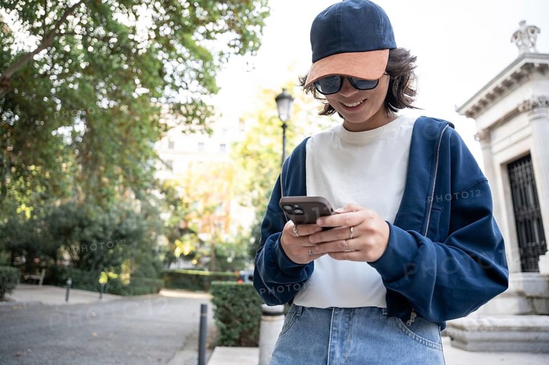 A young person smiles while looking at their phone during a leisurely walk. Dressed casually, they wear a cap and sunglasses against a background of trees and architectural features