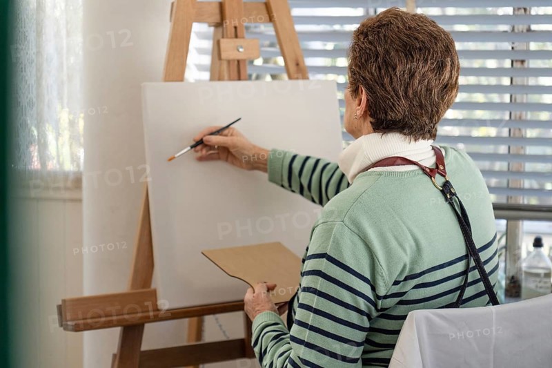 Rear view of a mature woman in a striped sweater holding a wooden palette while painting on a large white canvas on an easel in a bright home studio near a lace-curtained window