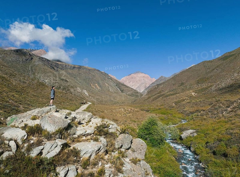 A lone male traveler stands on a rock, gazing over a serene stream winding through the lush valleys of the Andes, under a clear blue sky