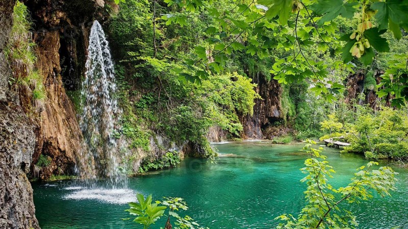 An enchanting waterfall pours into a turquoise river surrounded by lush foliage and rocks, Plitvice Lakes National Park, Croatia
