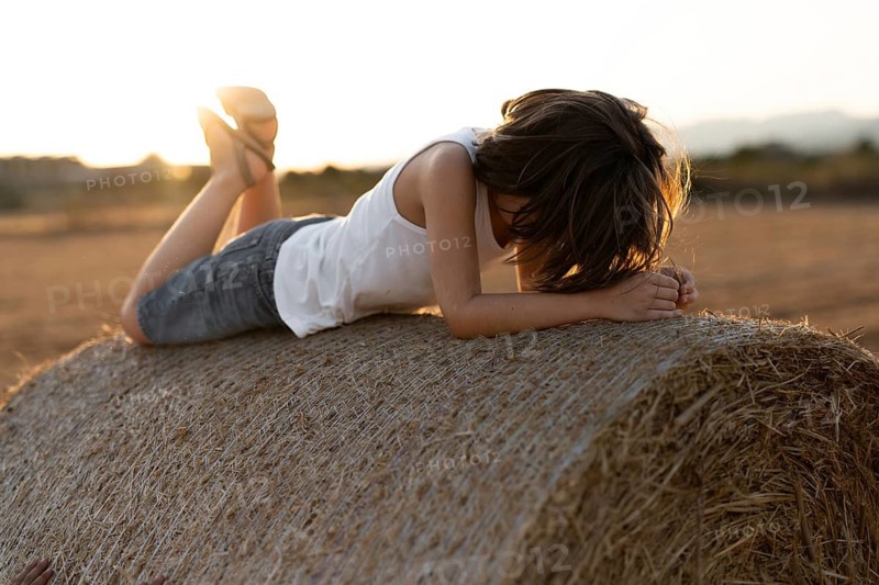 A child in a white tank top and denim shorts lies on a hay bale, enjoying a serene sunset in the countryside. The warm light adds a gentle glow to the peaceful scene