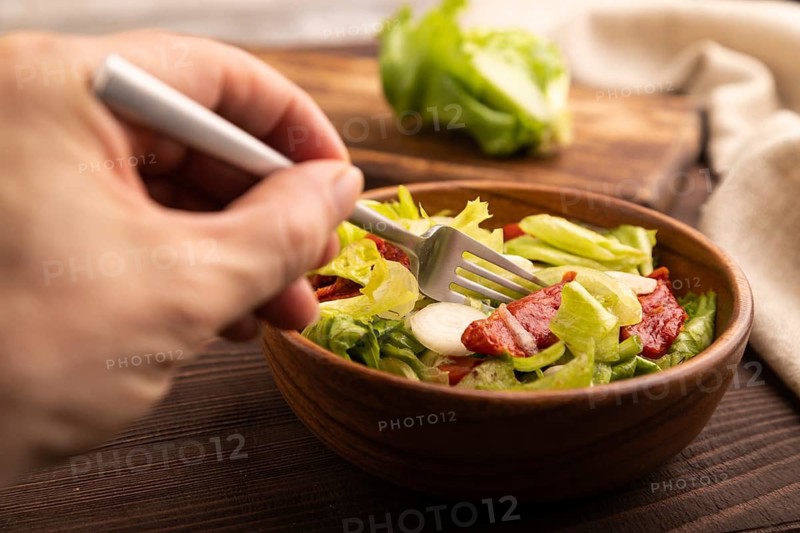 Mixed salad with lettuce, tomatoes, onion and vegetables in wooden bowl with hand on brown wooden background and linen textile. side view, close up, selective focus