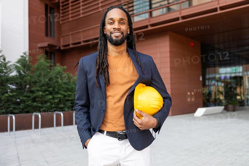 A cuban businessman stands confidently outside a modern building, holding a yellow hard hat. He is dressed in a suit, representing expertise and professionalism in the construction industry