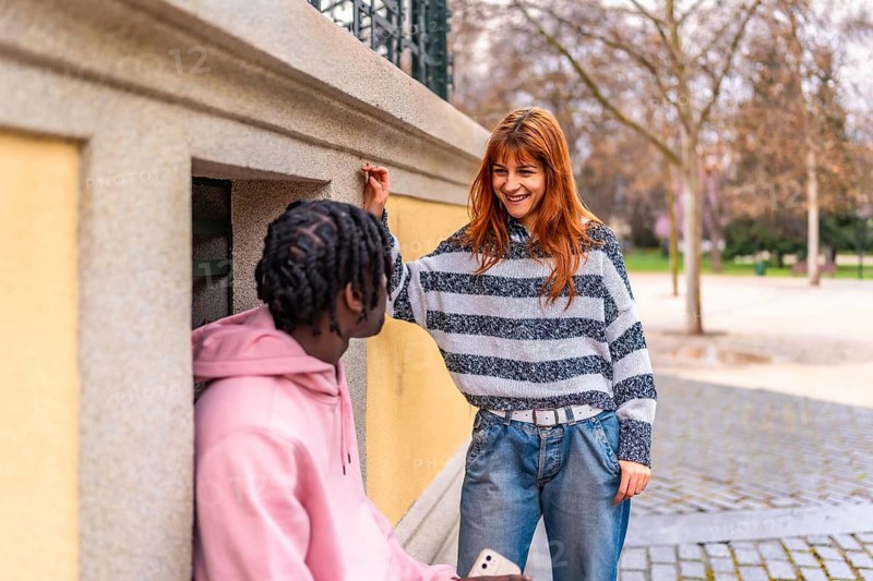Young woman with long red hair is leaning against a wall, laughing and talking to a friend in a park