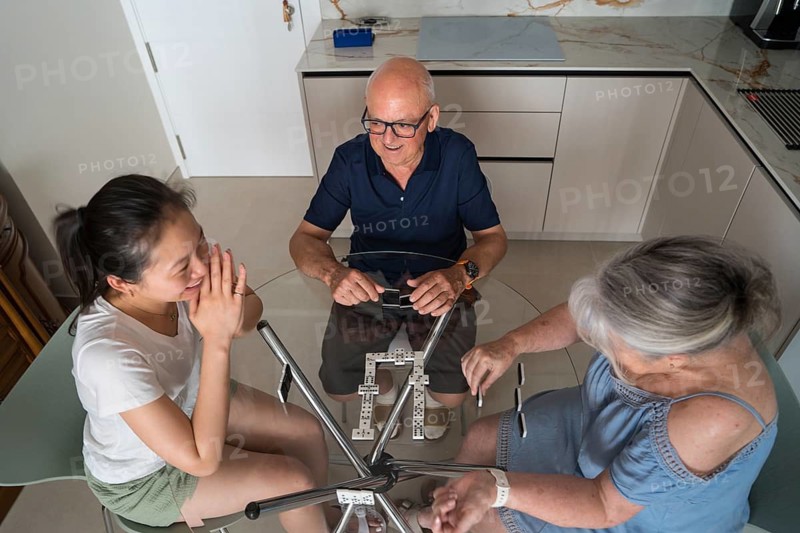 A joyous multigenerational family gathers around a glass kitchen table, deeply immersed in a lively game of dominoes, sharing laughter and creating cherished memories together