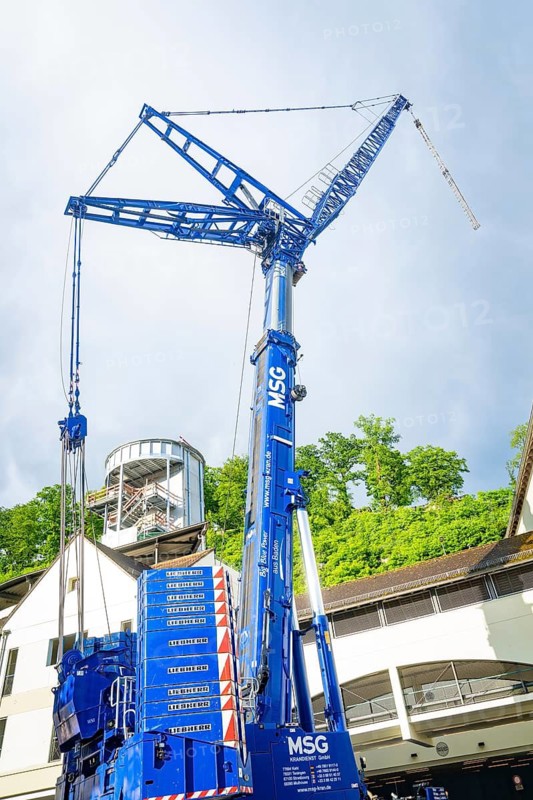 A large blue construction crane in front of a modern building under a cloudy sky, Hermann Hesse railway bridge lift, Calw, Germany