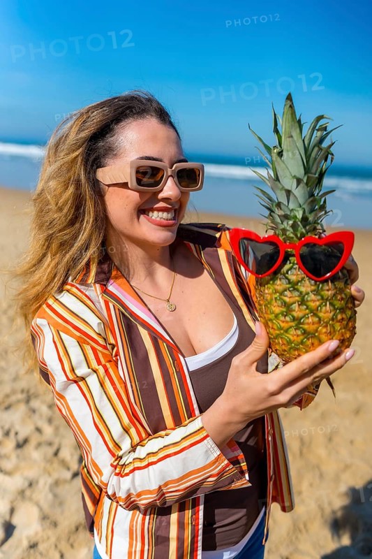 Woman wearing sunglasses and a striped shirt smiling while holding a fresh pineapple adorned with red heart shaped sunglasses, enjoying a tropical summer vacation by the ocean