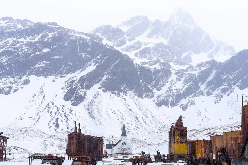 Norwegian wooden church between rusted industrial plants of the former whaling station, snow-capped mountains in the background during snowfall, Grytviken, South Georgia