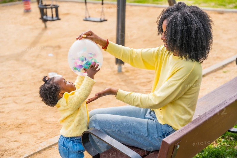 African mother and daughter playing with a balloon sitting on a park bench outdoors