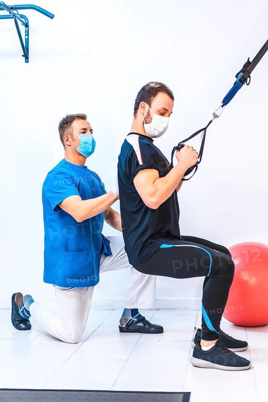 Physiotherapist in blue gown and a patient with rubber arm exercises. Physiotherapy with protective measures for the Coronavirus pandemic, COVID-19. Osteopathy, sports chiromassage