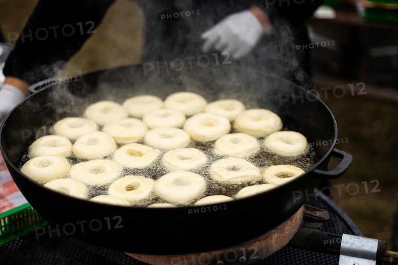 Cropped unrecognizable person cooking doughnuts in a large, black frying pan filled with oil at a bustling street fair. The scene captures steaming hot, fresh doughnuts immersed in sizzling oil, highlighting the vibrant and traditional aspect of street food culture