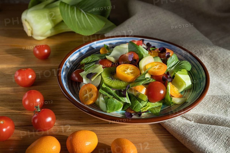 Vegetarian salad of pac choi cabbage, kiwi, tomatoes, kumquat, microgreen sprouts on a wooden background and linen textile. Hard light, contrast. Side view, close up