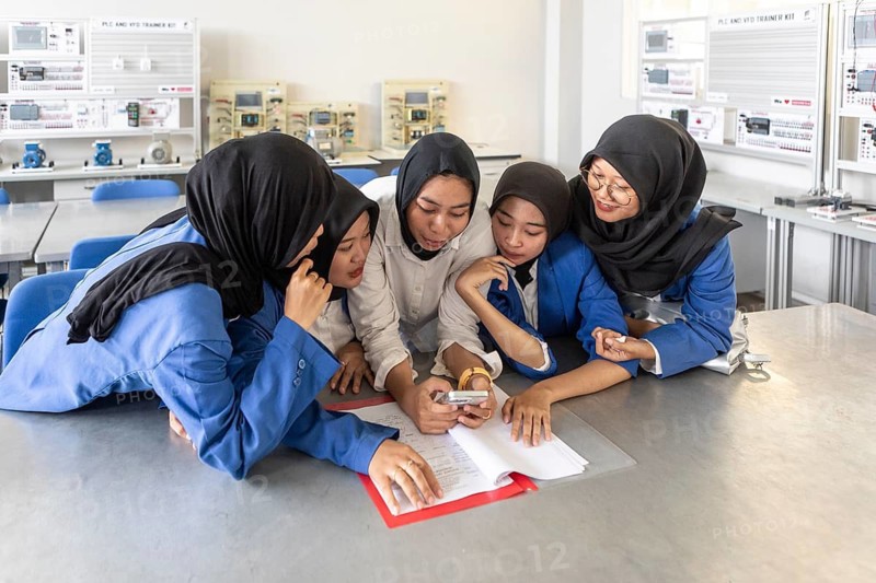 A group of Asian Muslim women in hijabs enthusiastically study together in a classroom, sharing a smartphone while reviewing papers and documents on the table