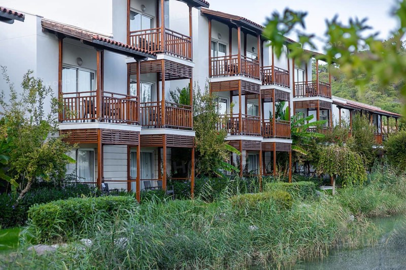 A modern building with wooden balconies surrounded by greenery. A hammock is visible in the garden area, creating a relaxing atmosphere