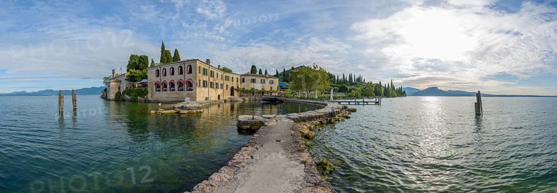 A serene view of a historic lakeside building by a calm lake with a pier extending into the water, surrounded by trees and mountains under a clear sky, Locanda San Vigilio, Punta San Vigilio, Garda, Lake garda, Lago di Garda, Veneto, Italy