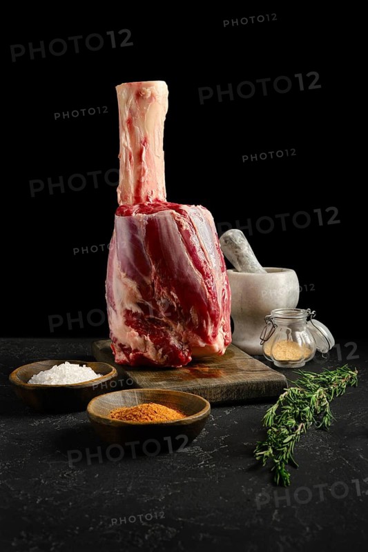 A large cut of beef hindshank on a wooden board with salt, spices, and herbs nearby. A mortar and jar with dry garlic on background