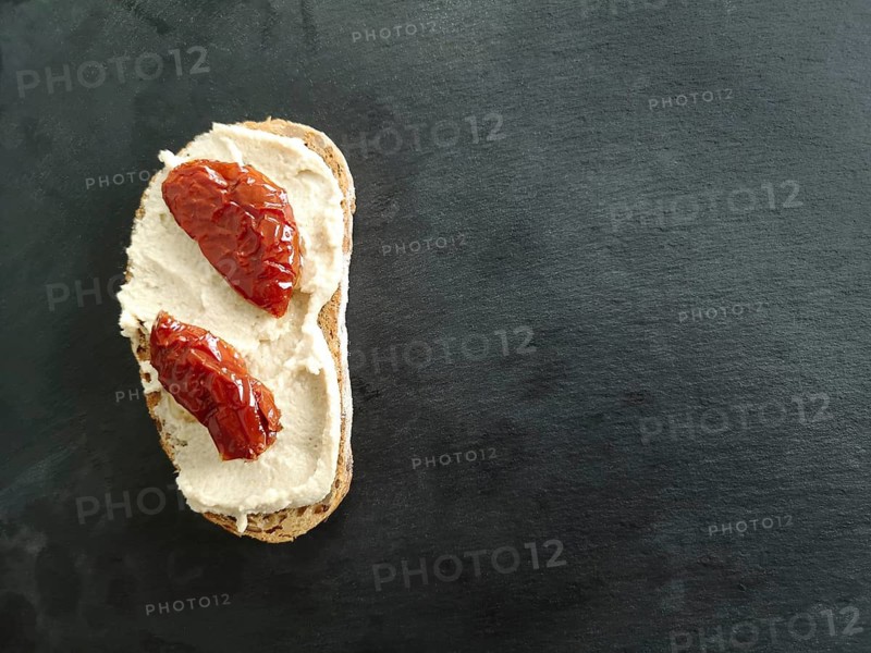A piece of bread with soft cheese and Sun-dried tomato on a table of dark grey stone slate, view from above