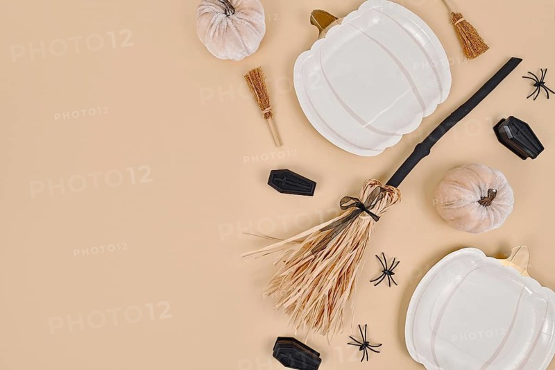 Halloween party flat lay with pumpkin shaped plates, witch broom, spiders and coffins on beige background with copy space