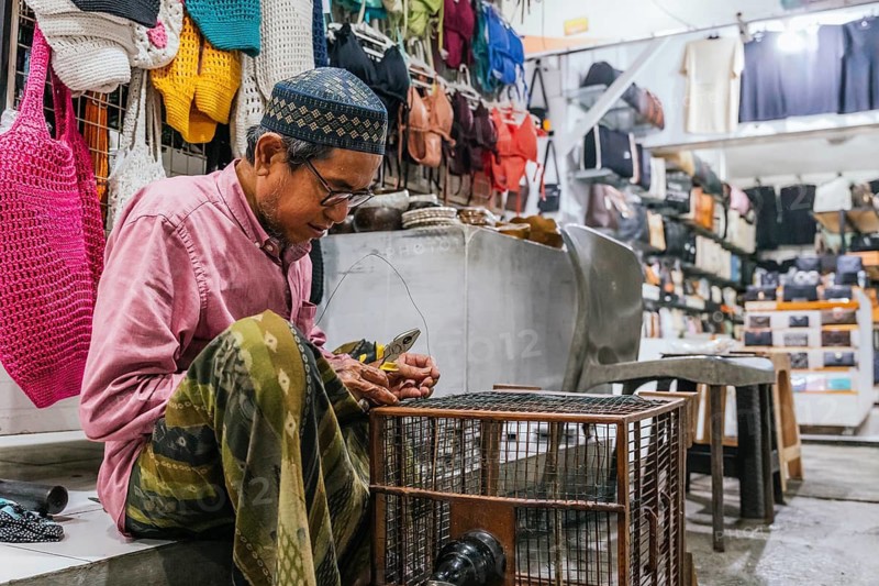 A balinese artisan focuses on his craft in a lively market. He works intently with tools near a colorful array of handmade bags and textiles, showcasing traditional techniques