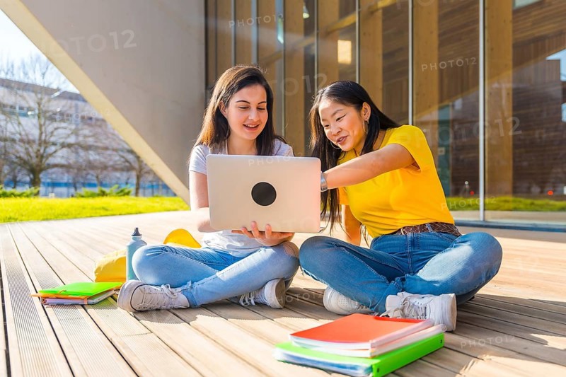 Asian and caucasian students using laptop in the campus sitting next to the university building