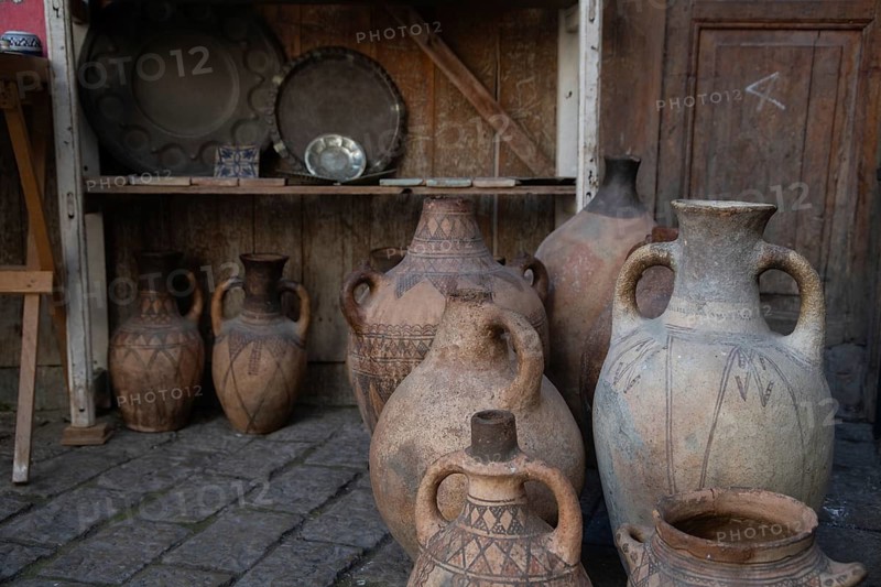 A collection of ancient clay jugs and pots displayed on a cobblestone floor in Fez, Morocco. The pottery features intricate patterns and earthy tones, highlighting historical craftsmanship