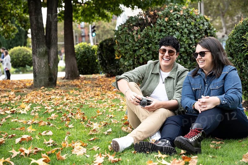 A joyful lesbian couple sits amidst autumn leaves in a park, sharing laughter. With casual attire and sunglasses, they capture the essence of a peaceful day outdoors