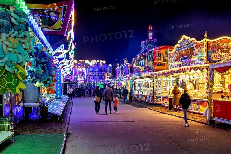 An illuminated fairground street at night with people walking along colorful stalls, carousel and the hustle and bustle of summer winnings in Eisenach