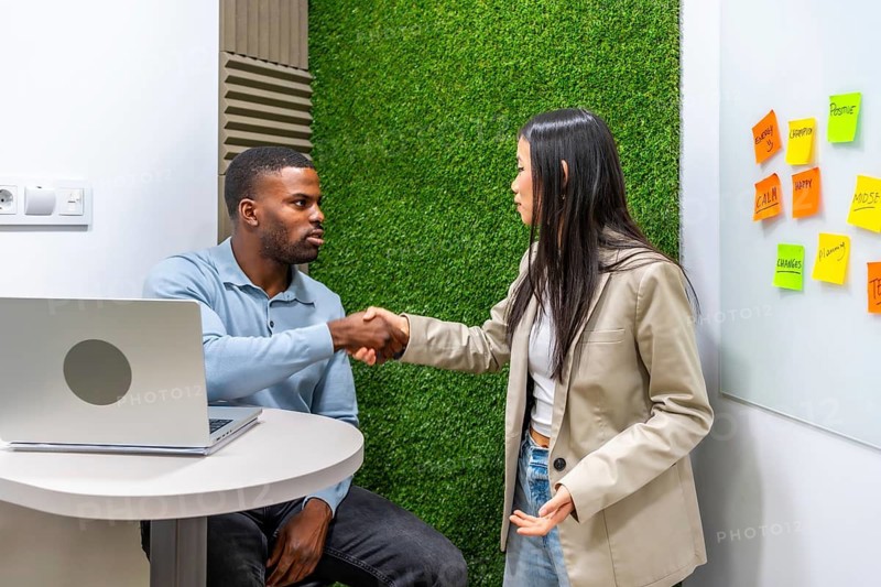 Two businesspeople shaking hands after reaching an agreement in a modern coworking office with a green wall and sticky notes