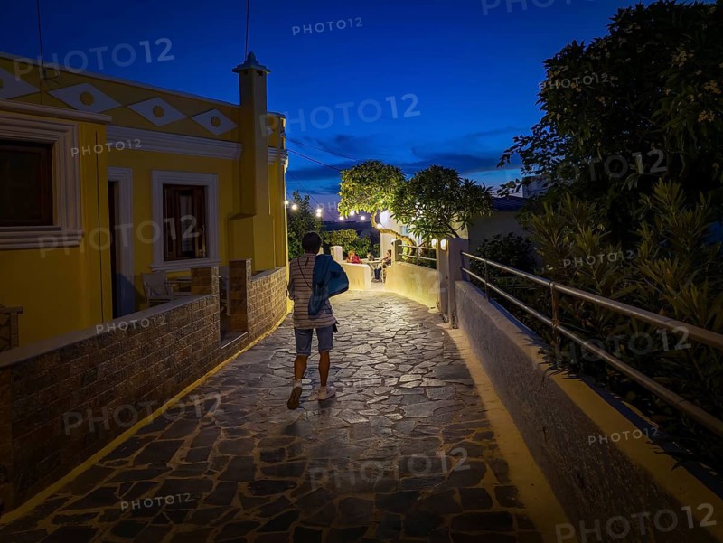 Evening mood of the illuminated promenade of Arkasa, yellow house and strollers in a cosy atmosphere, Arkasa, Karpathos, Greece