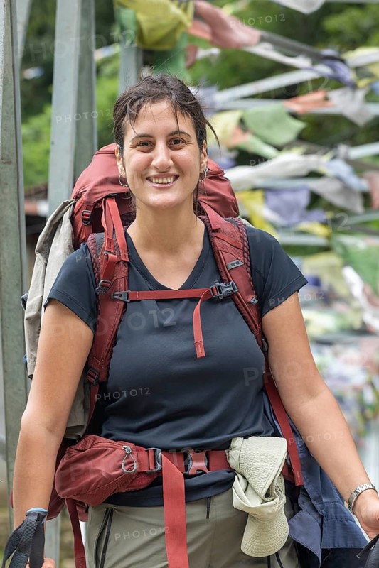 A woman smiles broadly as she embarks on a trek through the Everest region. Dressed in practical hiking attire and carrying a large backpack, she exudes enthusiasm and adventure