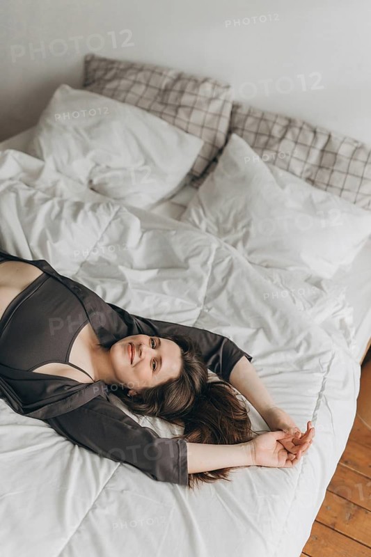 A cheerful young woman lies on a white bed with her arms stretched out, smiling and looking relaxed. The cozy setting and natural light create a warm and inviting atmosphere