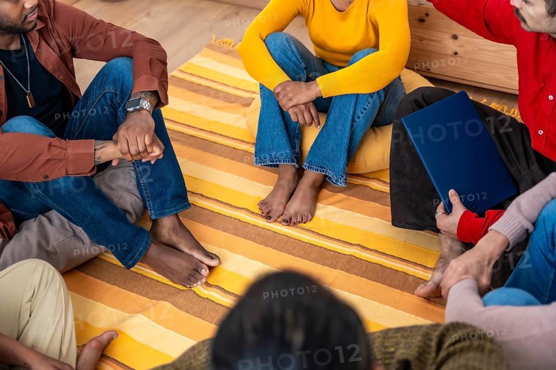 Diverse adults sit barefoot in a circle on an orange yellow rug, holding hands and listening attentively during a warm, informal group therapy session for mental health and support