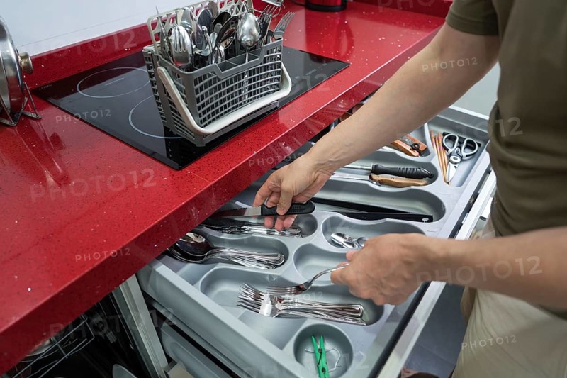 A person organizes utensils in a kitchen drawer, sorting knives, forks, and spoons in a modern home with a red countertop, emphasizing household order and cleanliness