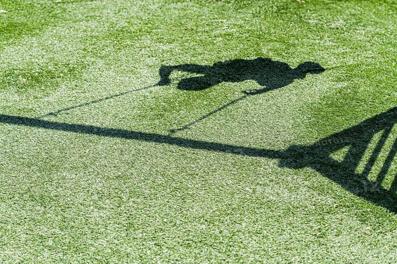 A shadow of a boy swinging joyfully on a playground swing is cast on lush green grass. The sunlit scene captures the essence of carefree childhood and outdoor fun