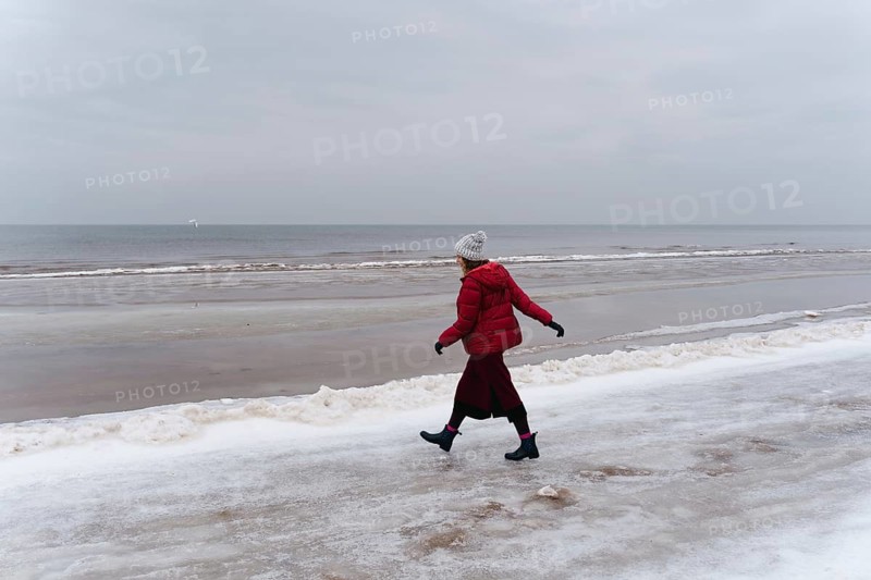 A woman in red walks in winter along the seashore, A person in a red coat walks alone along a snowy beach under a gray sky, Jurmala, Latvia