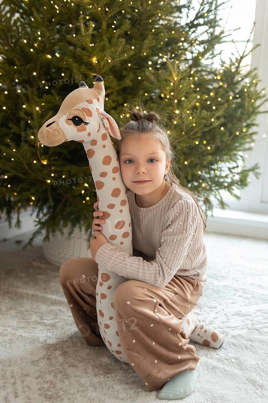 A young Caucasian girl looks directly at the camera while holding a stuffed giraffe in front of a brightly lit Christmas tree. She is dressed in a cozy ribbed sweater and pants with contrasting polka dots