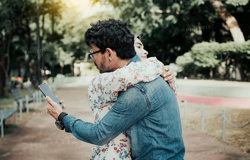 Unfaithful man hugging his girlfriend and looking at the cell phone. Concept of unfaithful man using cell phone. Unfaithful man looking at the cell phone while hugging his girlfriend outdoors