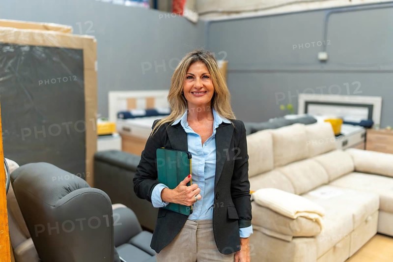Smiling mature businesswoman holding a tablet while standing in a furniture showroom, assisting customers and managing sales in a home improvement store