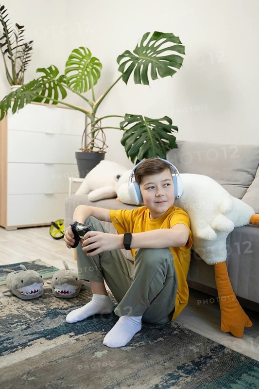 A boy wearing headphones sits on the floor playing video games with a controller in hand. Surrounded by plush toys and cozy home decor, he enjoys a relaxed moment in a modern living room