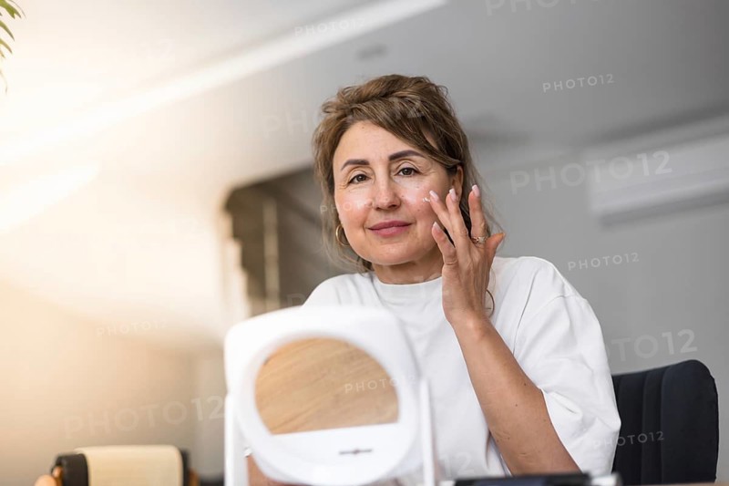 A woman is carefully applying moisturizer to her face while sitting at a table in a relaxed indoor setting. Natural light fills the room, highlighting her focused expression and self-care routine
