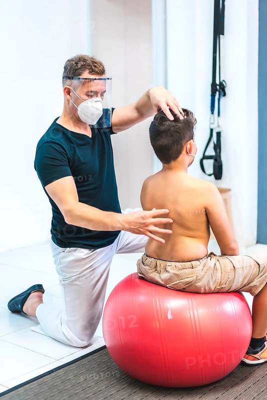 Physiotherapist with mask and screen looking at a child's back. Reopening with security measures of physiotherapists in the Covid-19 pandemic. Osteopathy, therapeutic chiromassage