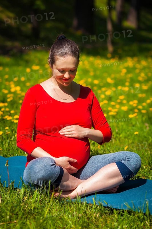 Pregnancy yoga exercise, pregnant woman doing asana Sukhasana easy yoga pose holding her abdomen outdoors on grass lawn with dandelions in summer