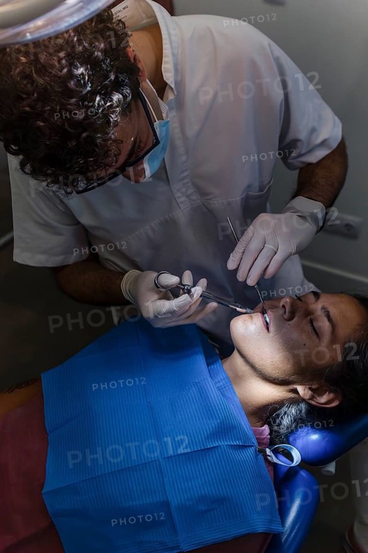 Dentist performing a teeth cleaning on a patient in a clinic setting. The patient is seated in a dental chair, and the dentist is focused on the treatment process