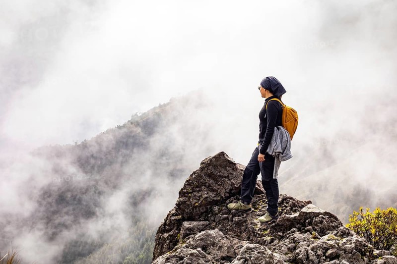 A hiker stands on a rocky peak, gazing over a mist covered mountain valley. Dressed in outdoor gear with a yellow backpack, they embrace the serenity and adventure of the moment