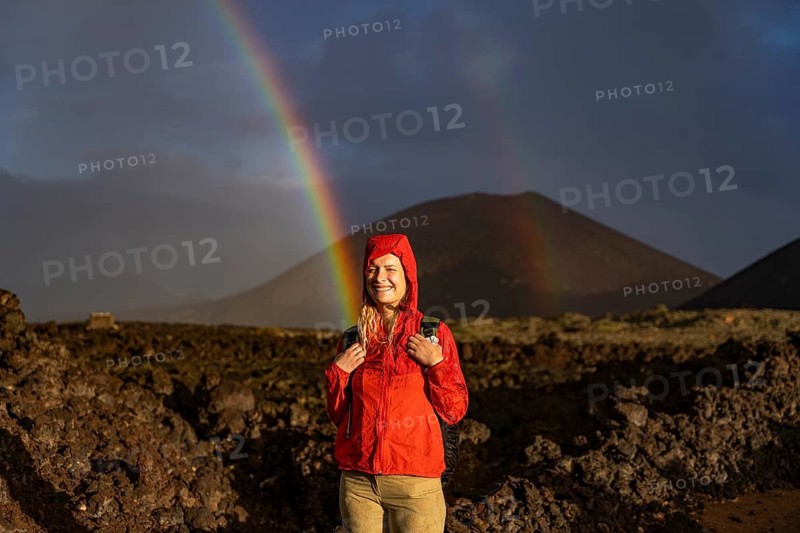 A joyful hiker in a bright red jacket stands in Timanfaya National Park, Lanzarote, with a vivid rainbow arching over volcanic terrain in the background