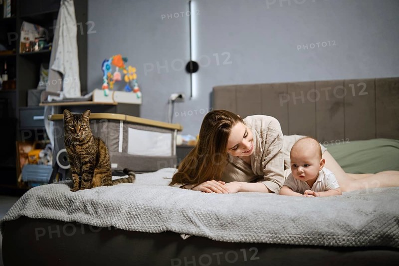 A mother lies on her stomach on a bed next to her infant, who is looking around. A cat sits nearby. The room is simple with soft light coming in. They share a moment of joy in their home