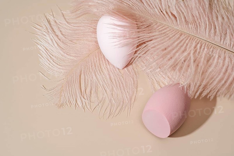 Aesthetic composition featuring two pink cosmetic sponges nestled on a soft beige surface surrounded by light pink feathers, evoking a sense of elegance and beauty
