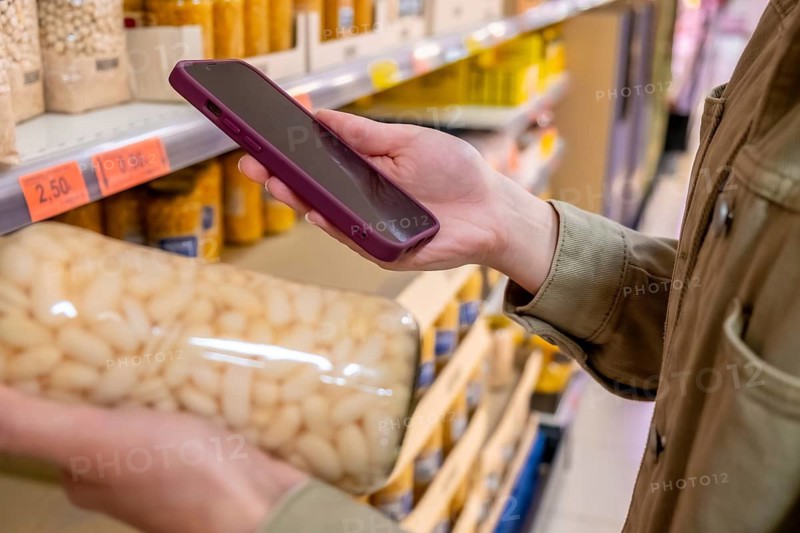 Person's hand holding a smartphone while checking a product on a supermarket shelf, using a mobile app to compare prices or manage a shopping list during grocery retail